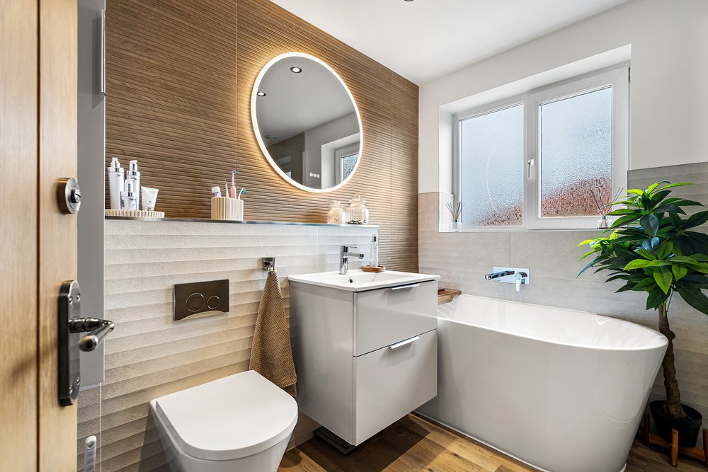 Contemporary bathroom with a wood-effect feature wall, backlit round mirror, wall-hung vanity unit and freestanding bath