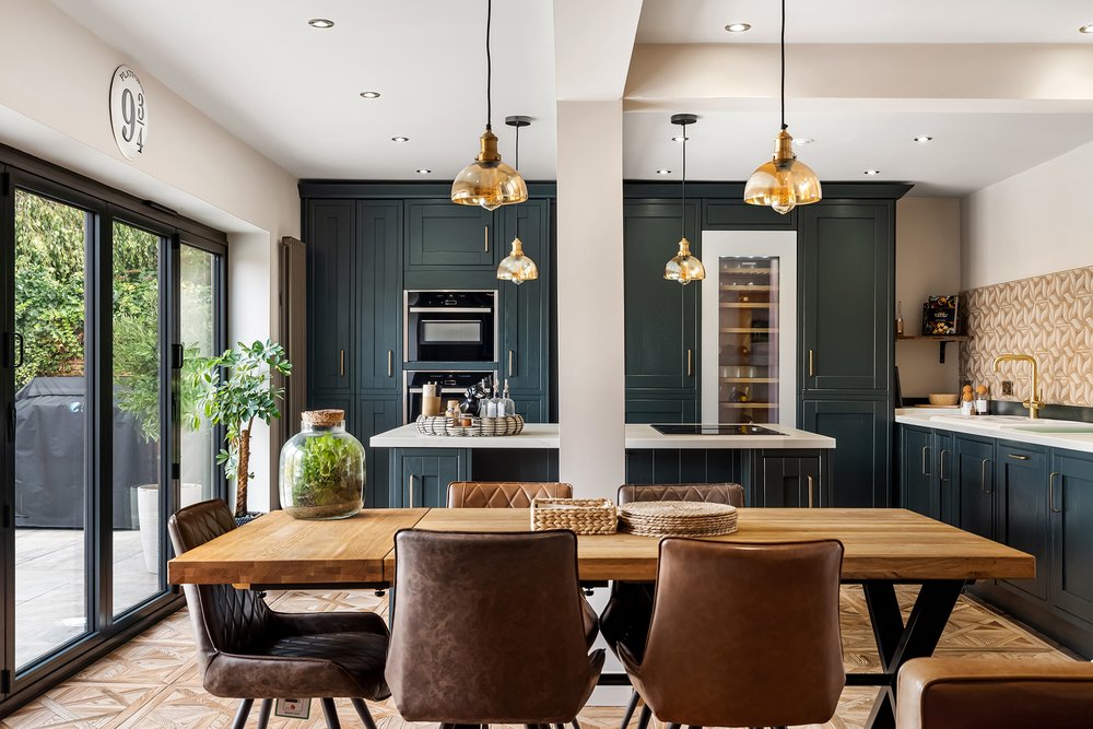 Stylish kitchen diner with dark green cabinetry, brass pendant lights, a wine cooler and an oak dining table beside sliding glass doors
